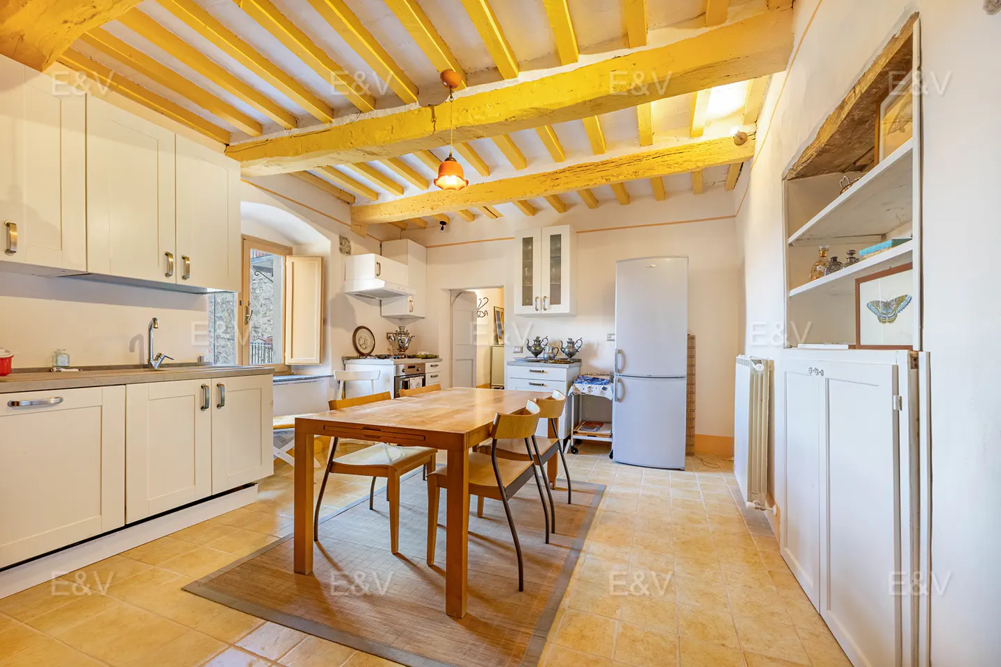 A bright kitchen with yellow beams, white cabinets, and a wooden table with chairs. A refrigerator and open shelving are visible.