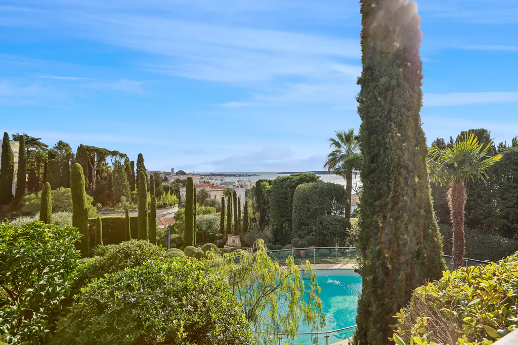 View of a blue pool surrounded by green trees, with a city and ocean in the background under a blue sky.