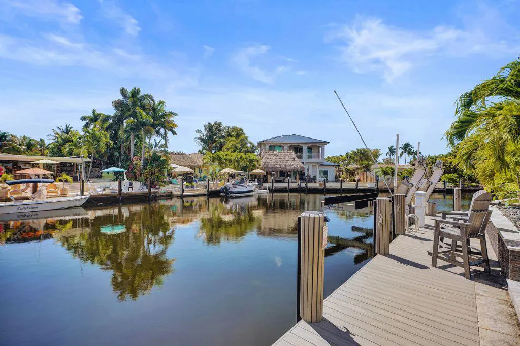 Waterfront property with a dock, boats, and palm trees under a blue sky. The water reflects the surrounding buildings and foliage.