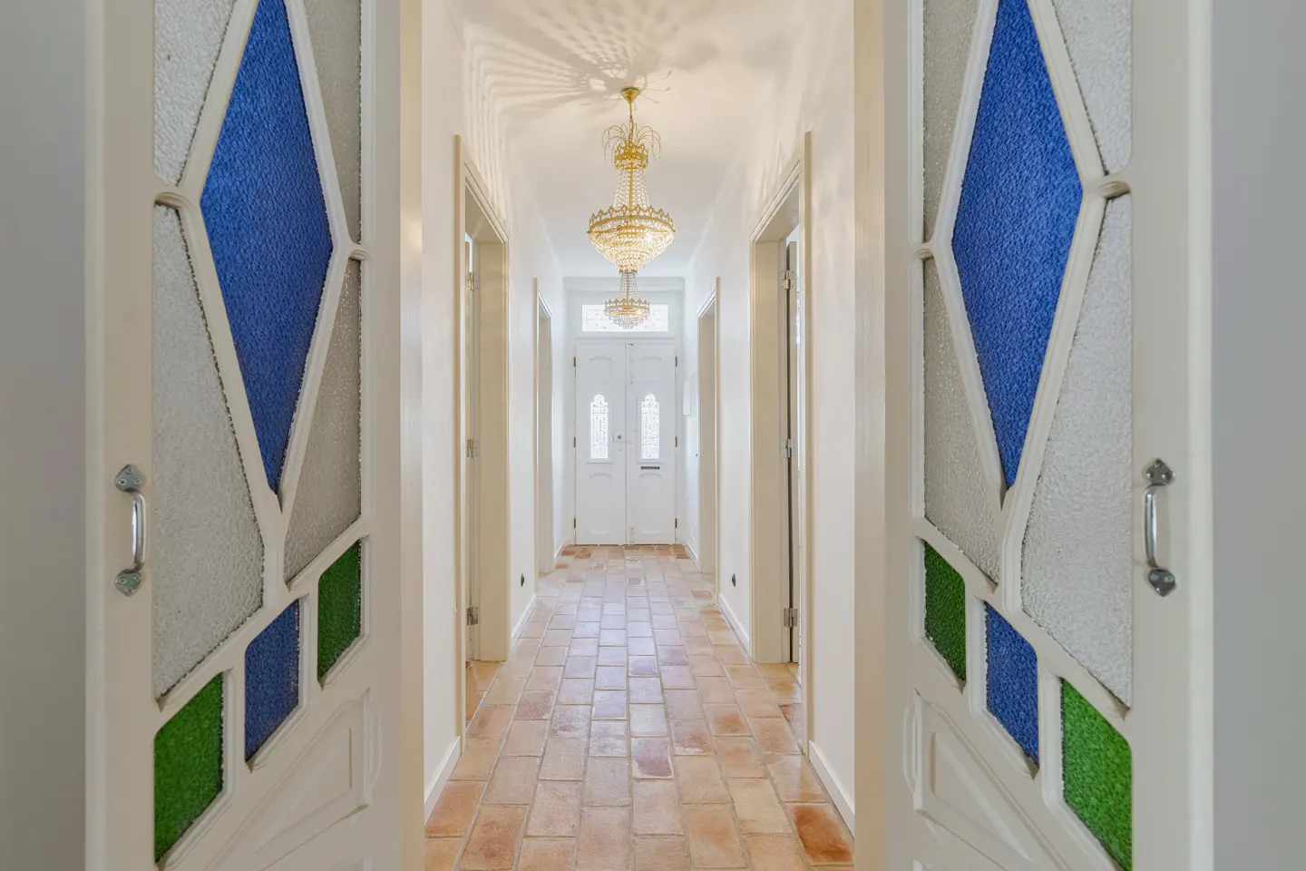 Hallway with terracotta tile floor, crystal chandelier, and white doors featuring blue and green stained glass accents.