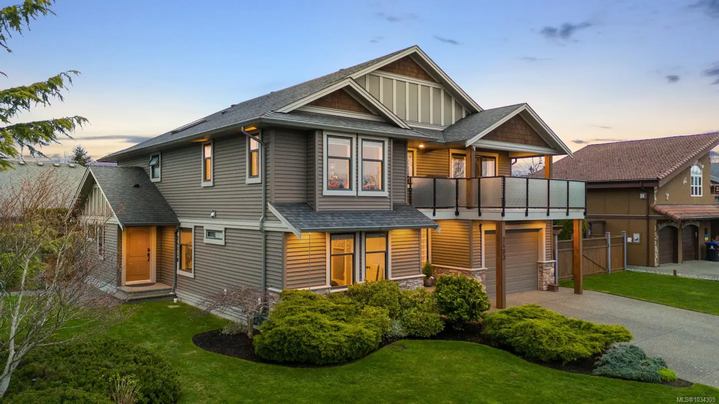 Two-story gray house with a balcony above the garage, green lawn, and blue sky.
