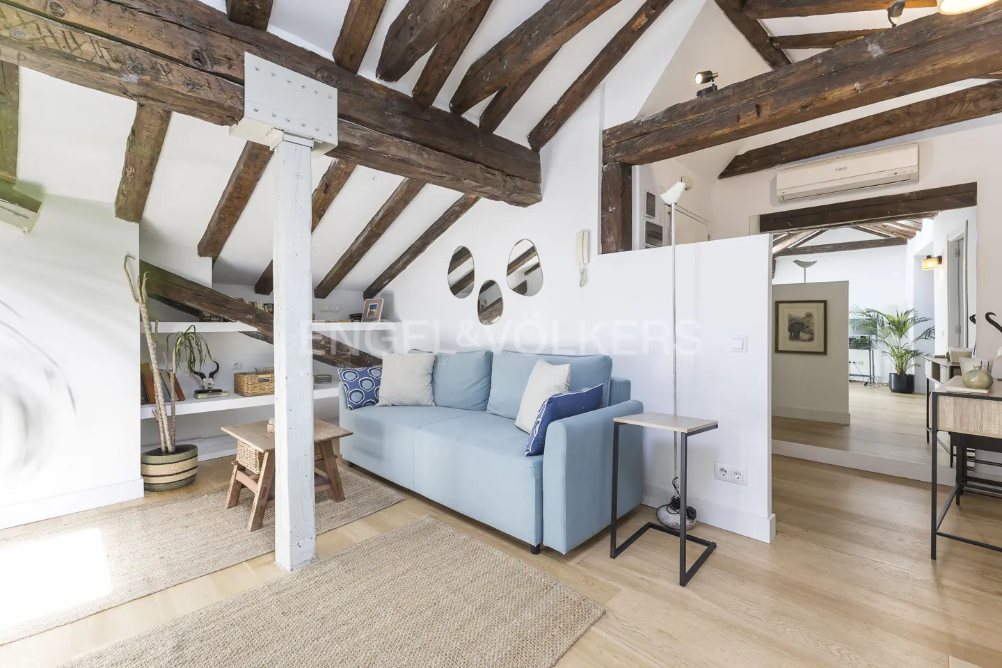 Living room with exposed wood beams, light blue sofa, and light wood floors. White walls and a partial wall divide the space.