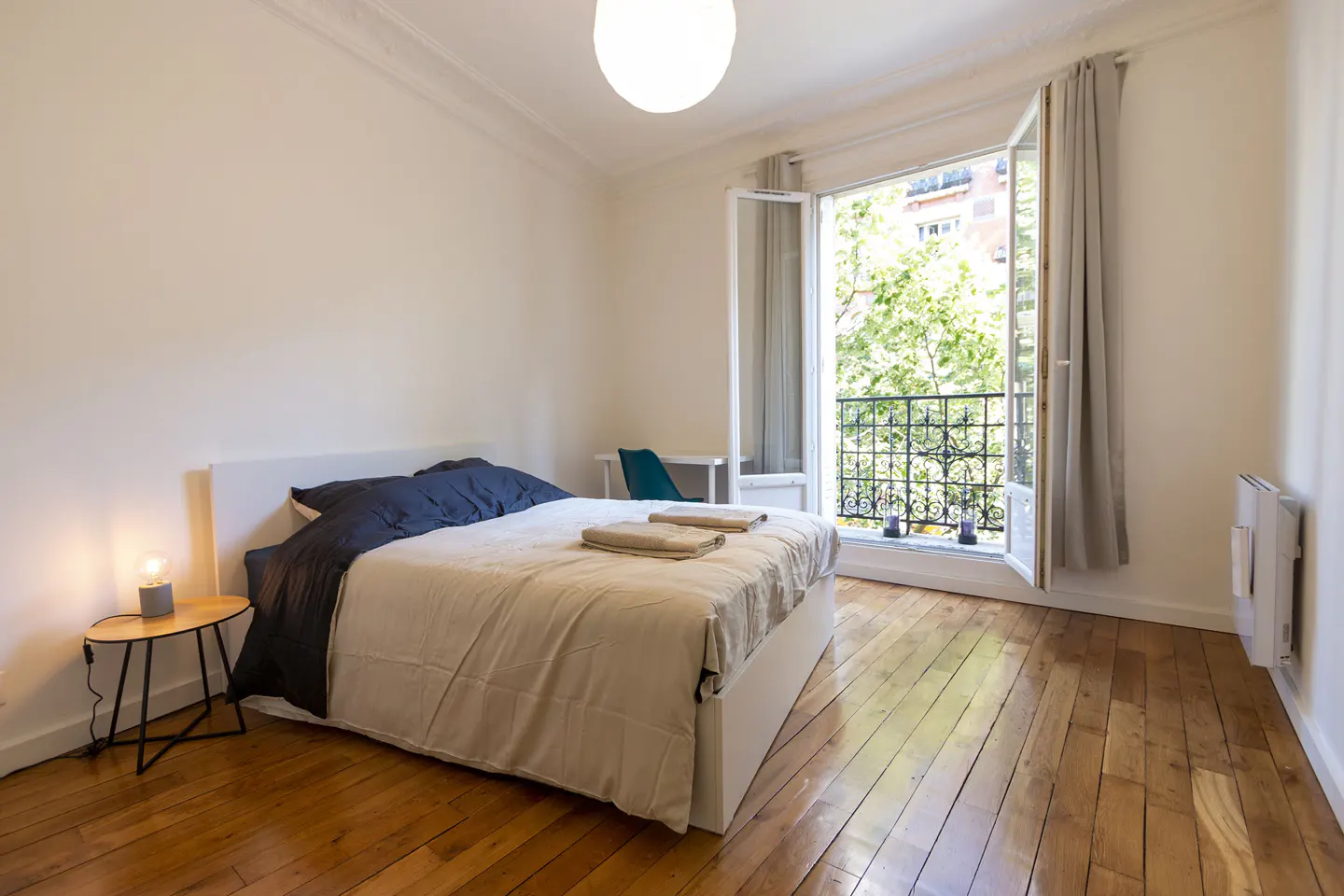 Bedroom with wood floors, a bed with blue sheets, and an open balcony door with a view of green trees.