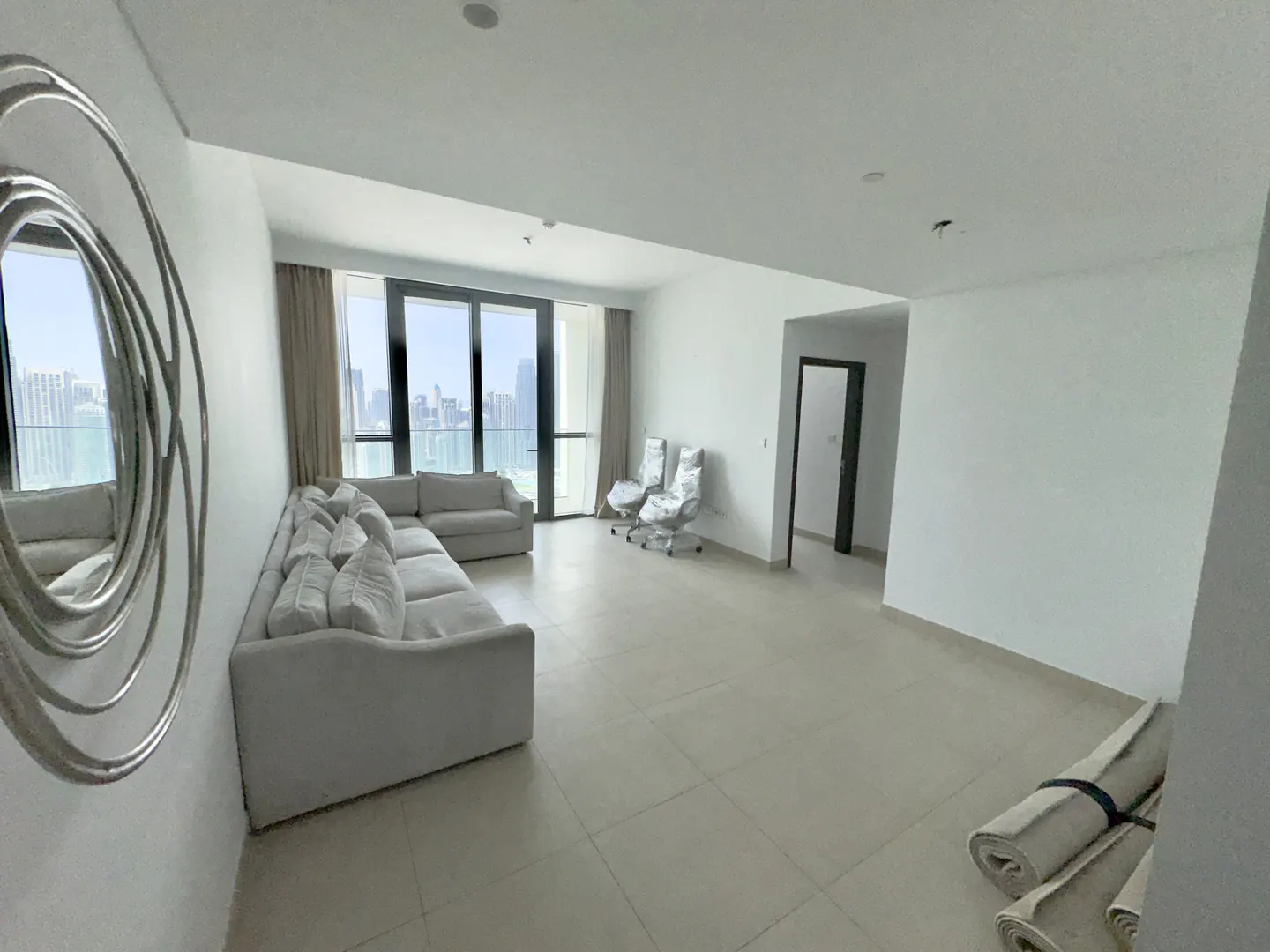 Bright, empty living room with a white sectional sofa, large windows, and a modern silver mirror on the wall. Two chairs are wrapped in plastic.
