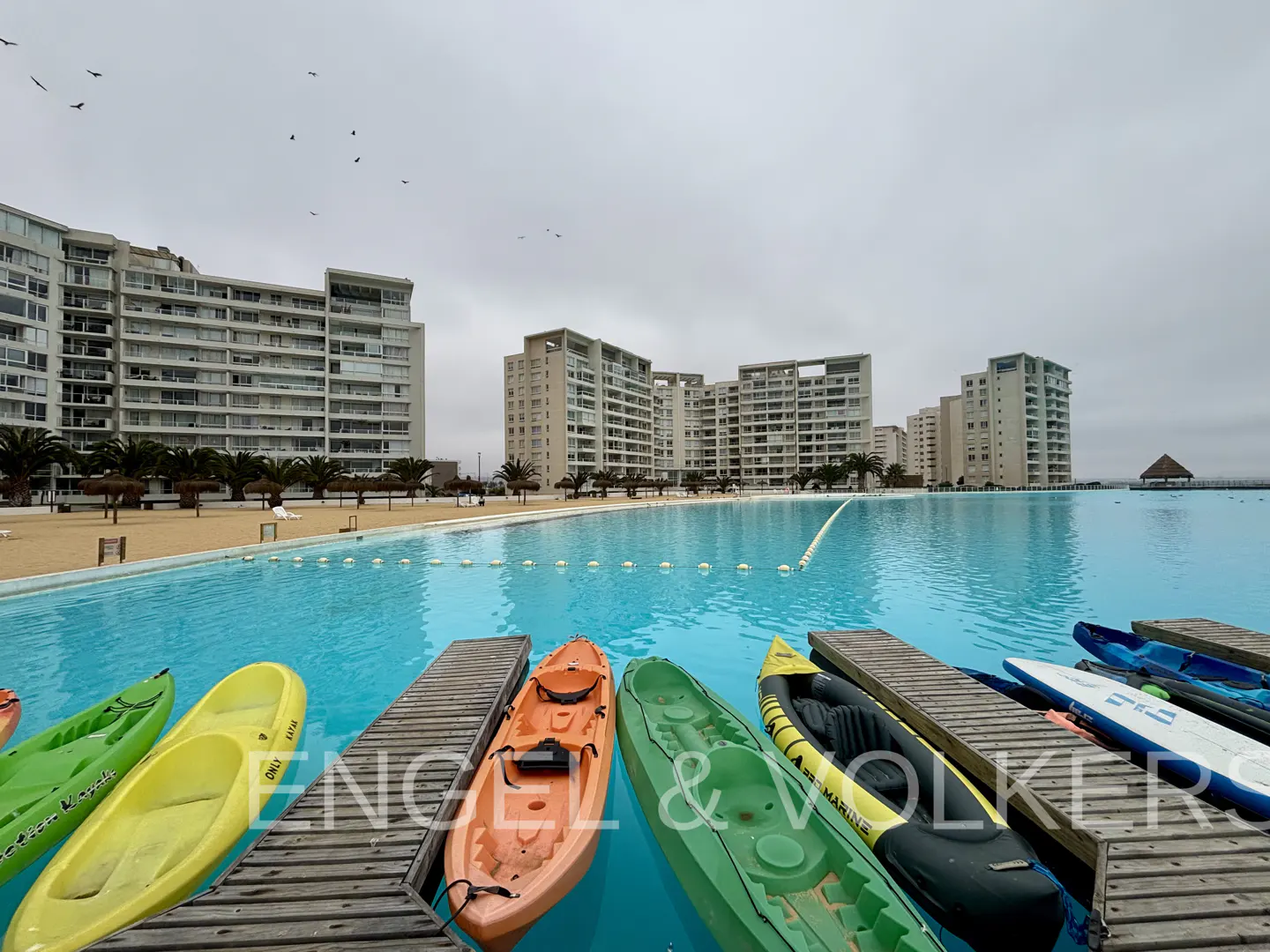 Kayaks and paddleboards on docks in a turquoise lagoon, with condos and palm trees in the background on a cloudy day.