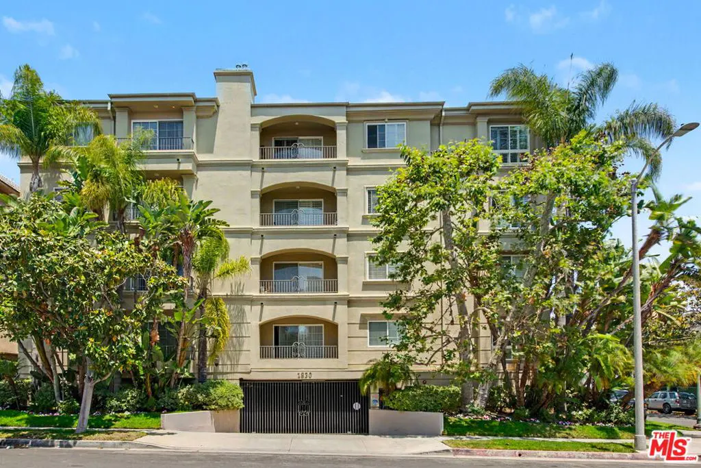 Exterior view of a four-story beige apartment building with balconies and a black gated entrance. Palm trees and green foliage surround the building.