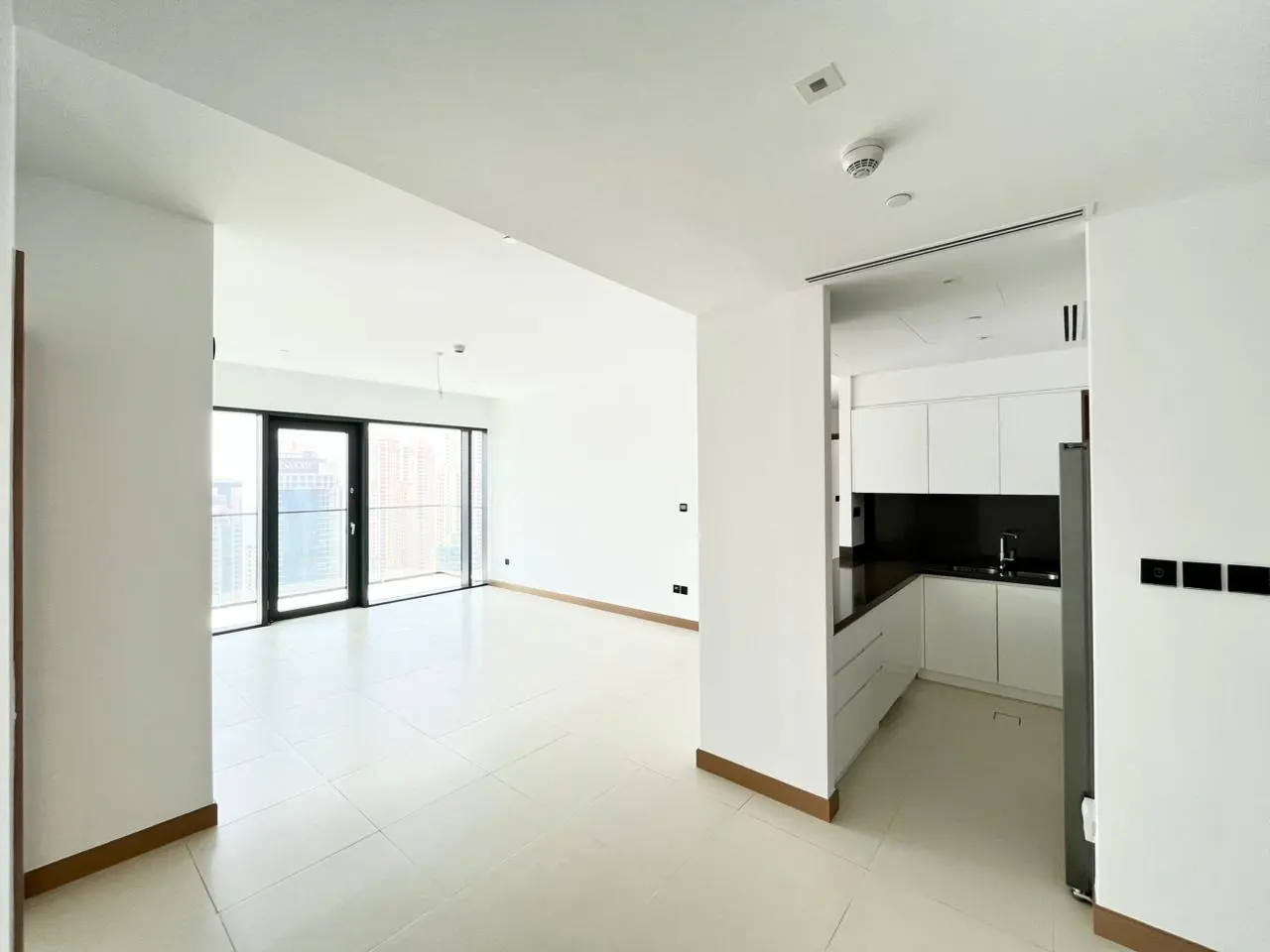 Bright, modern apartment with white walls, tile floors, and a black-framed window overlooking a city. A white kitchen is visible.