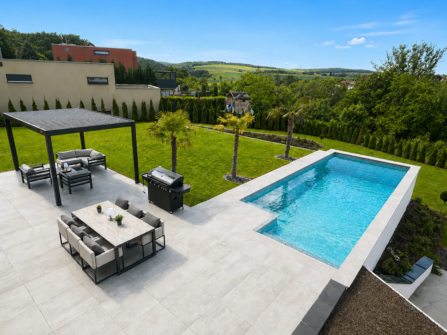 An aerial view of a backyard with a pool, patio furniture, grill, and green lawn. The sky is blue.