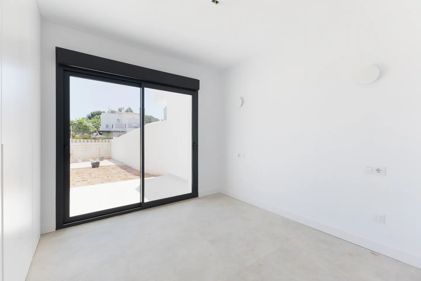 Minimalist room with white walls, light tile floors, and a black sliding glass door opening to a sunny outdoor patio.