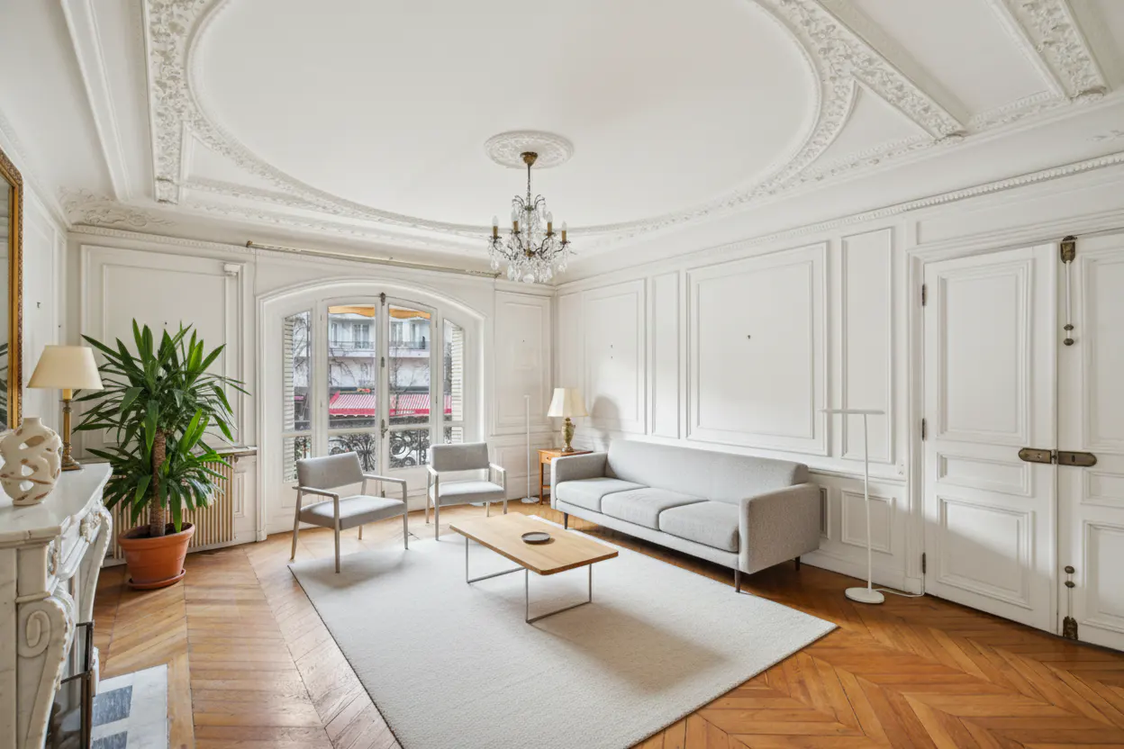 Bright living room with white walls, herringbone wood floors, and a large window. A gray sofa and chairs sit on a white rug. A chandelier hangs from the ornate ceiling.