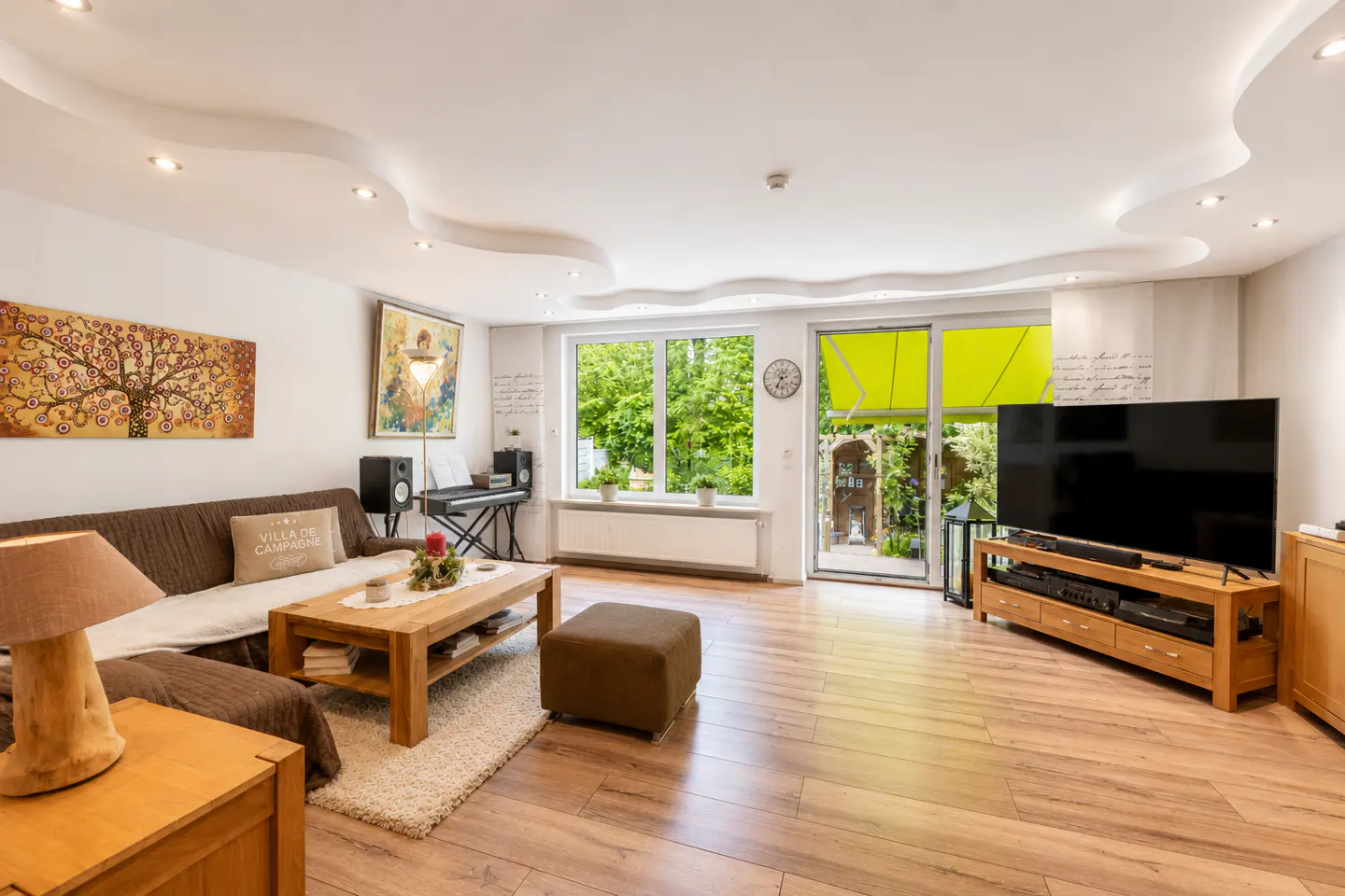 A bright living room with wood floors, a brown sofa, a TV, and a view of a green garden.
