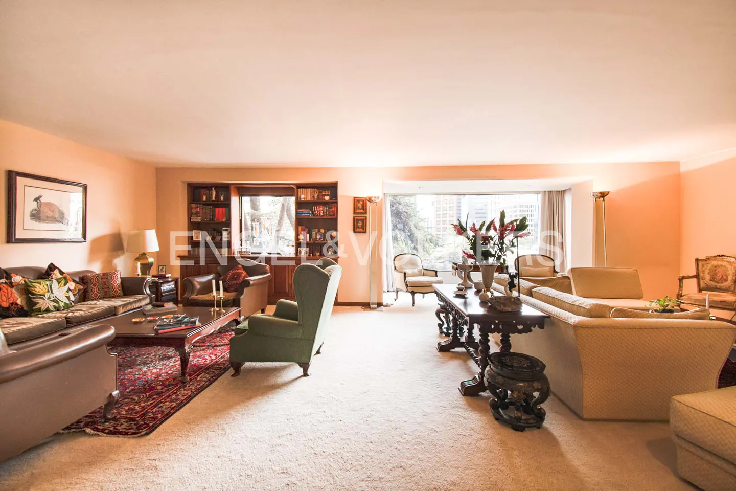 A wide shot of a living room with sofas, chairs, and a large window. The room is decorated with books, flowers, and artwork.