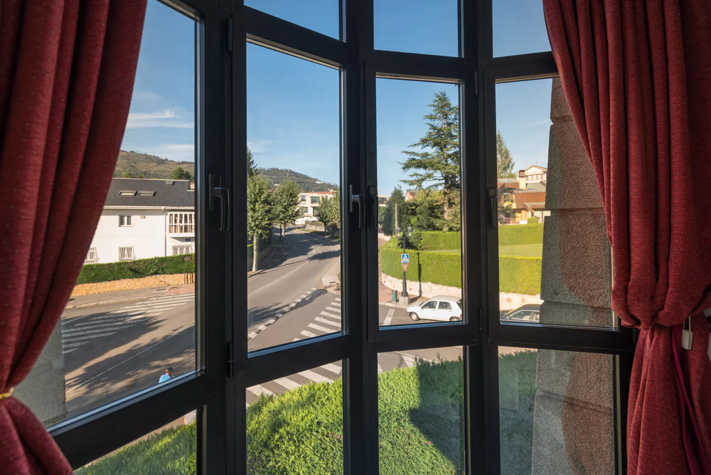 View from a window with red curtains, showing a street with houses, trees, and a white car on a sunny day.