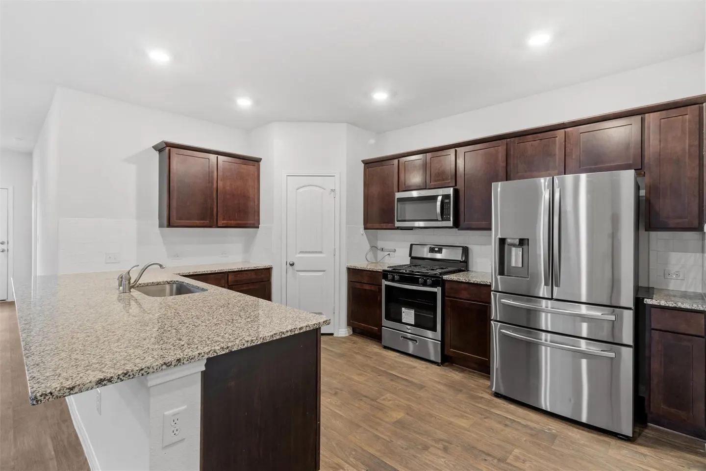 Bright kitchen with wood floors, white walls, and dark wood cabinets. Stainless steel appliances include a refrigerator, oven, and microwave. A granite countertop island has a sink.