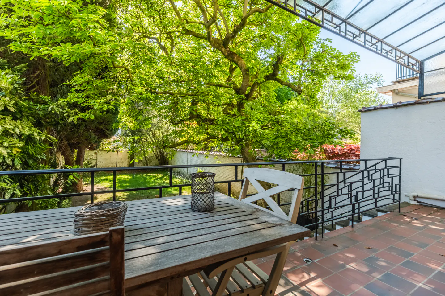 Outdoor patio with a wooden table, chairs, and a view of a lush green garden. Black metal railing and a clear roof structure overhead.
