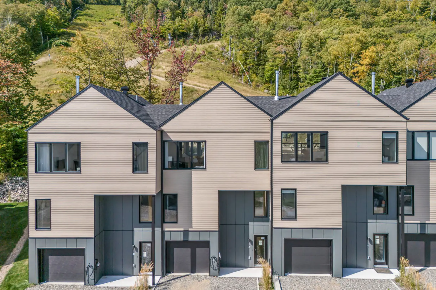 A row of modern townhouses with beige siding, dark gray accents, and black roofs, set against a backdrop of forested hills.
