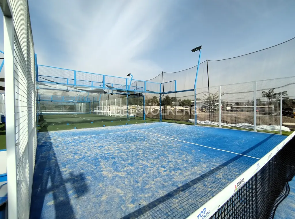 Outdoor blue paddle tennis court with netting, lights, and a blue sky.