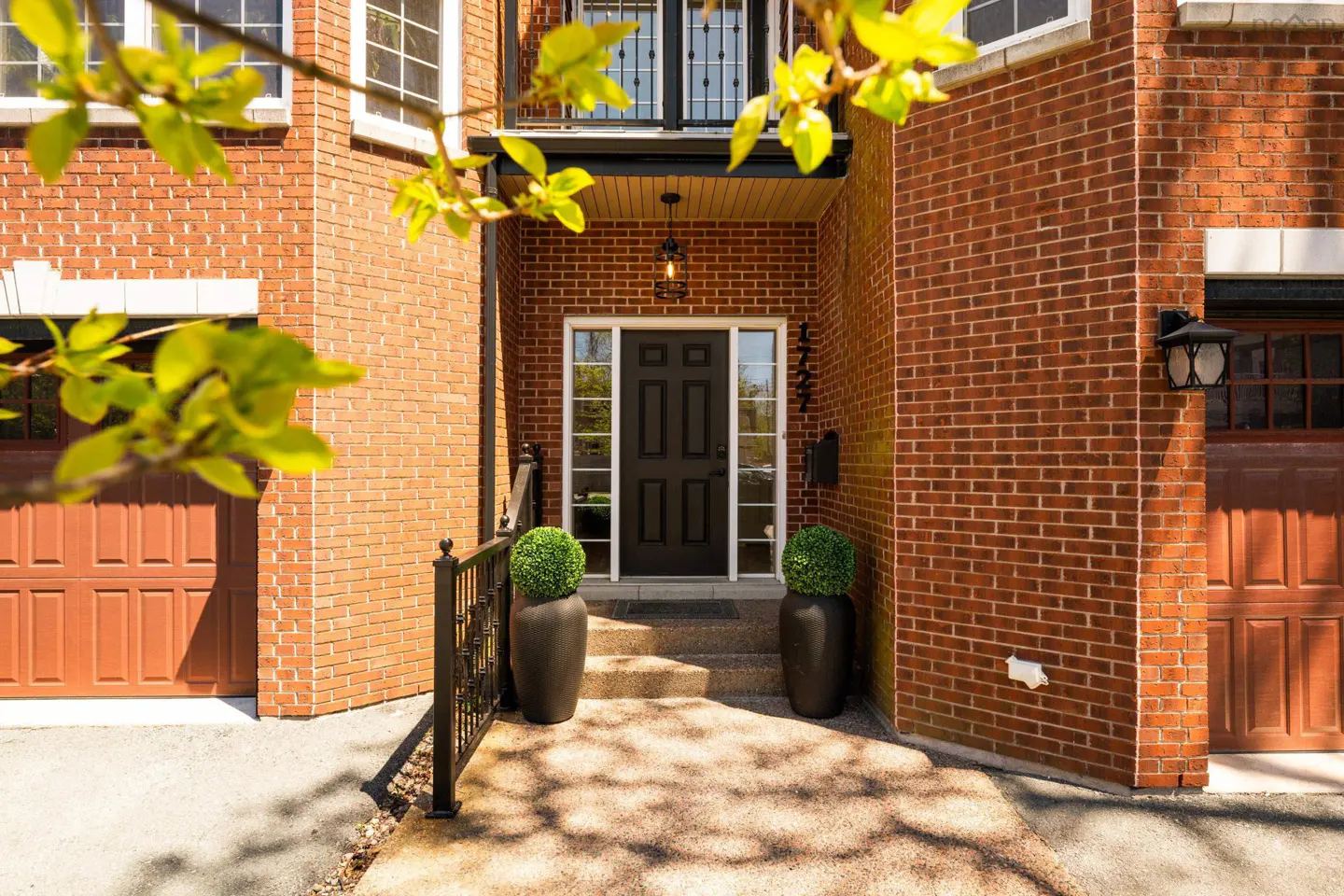 Front entrance of a red brick house with a black door, two potted topiary trees, and a garage on each side.