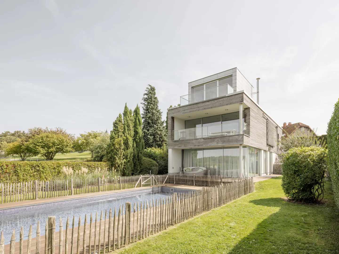 Modern three-story house with a pool. The house has large windows and balconies. A wooden fence surrounds the pool and yard.