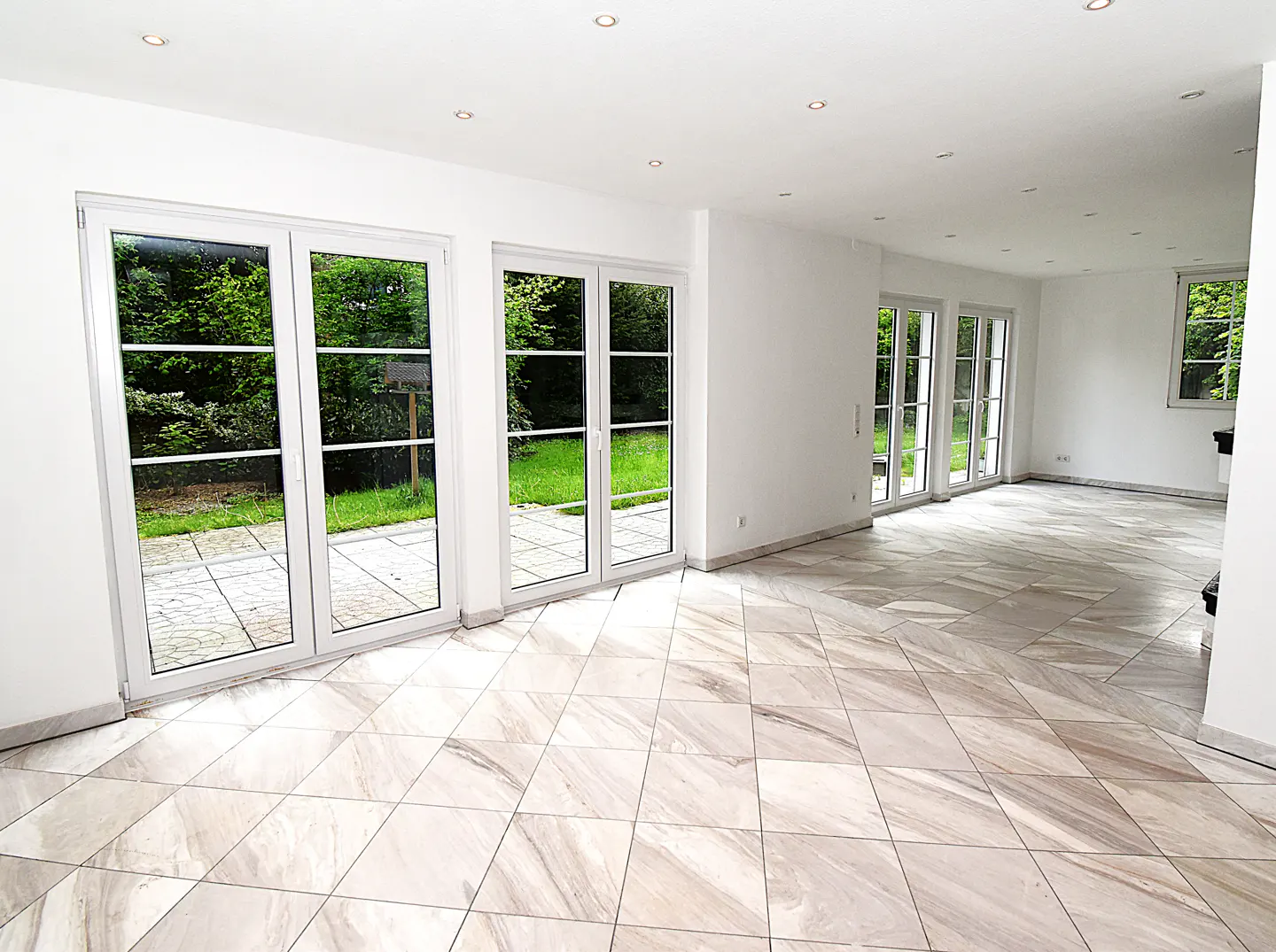 Bright, empty room with white walls, recessed lighting, and marble-patterned tile floor. Large windows offer a view of a green yard.