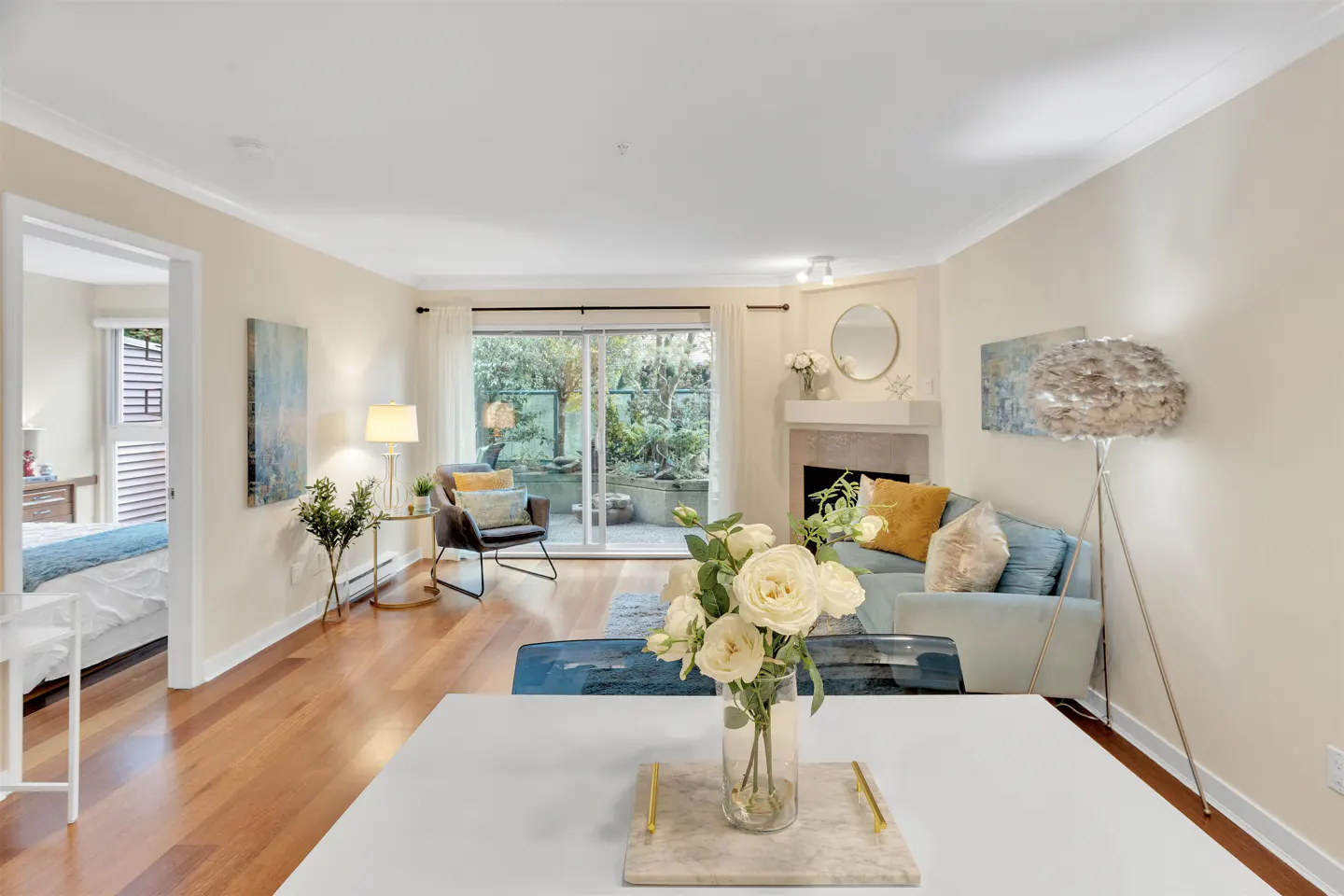 Bright living room with hardwood floors, a blue sofa, and a fireplace. A white dining table with flowers sits in the foreground. Patio doors lead to a garden.