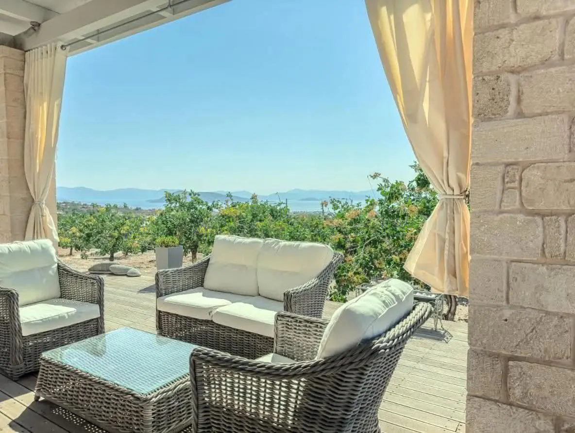Outdoor patio with wicker furniture, white cushions, and glass table. Ocean view with trees and blue sky. Cream curtains frame the scene.