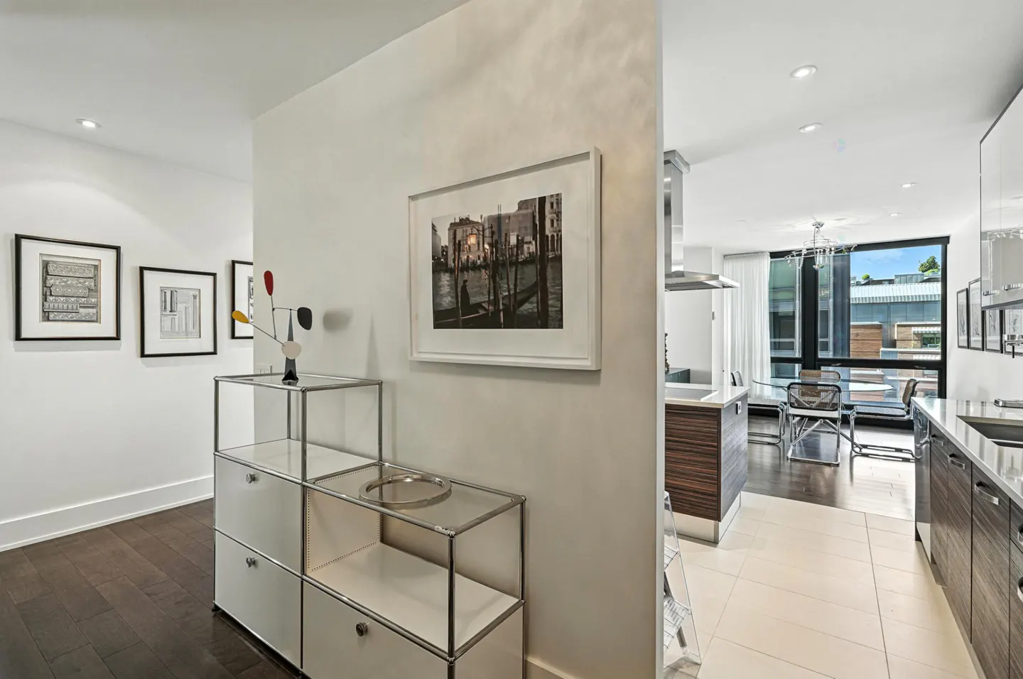 Hallway view into a modern kitchen. White walls, dark wood floors, and framed art. A white cabinet with a metal frame holds a sculpture.