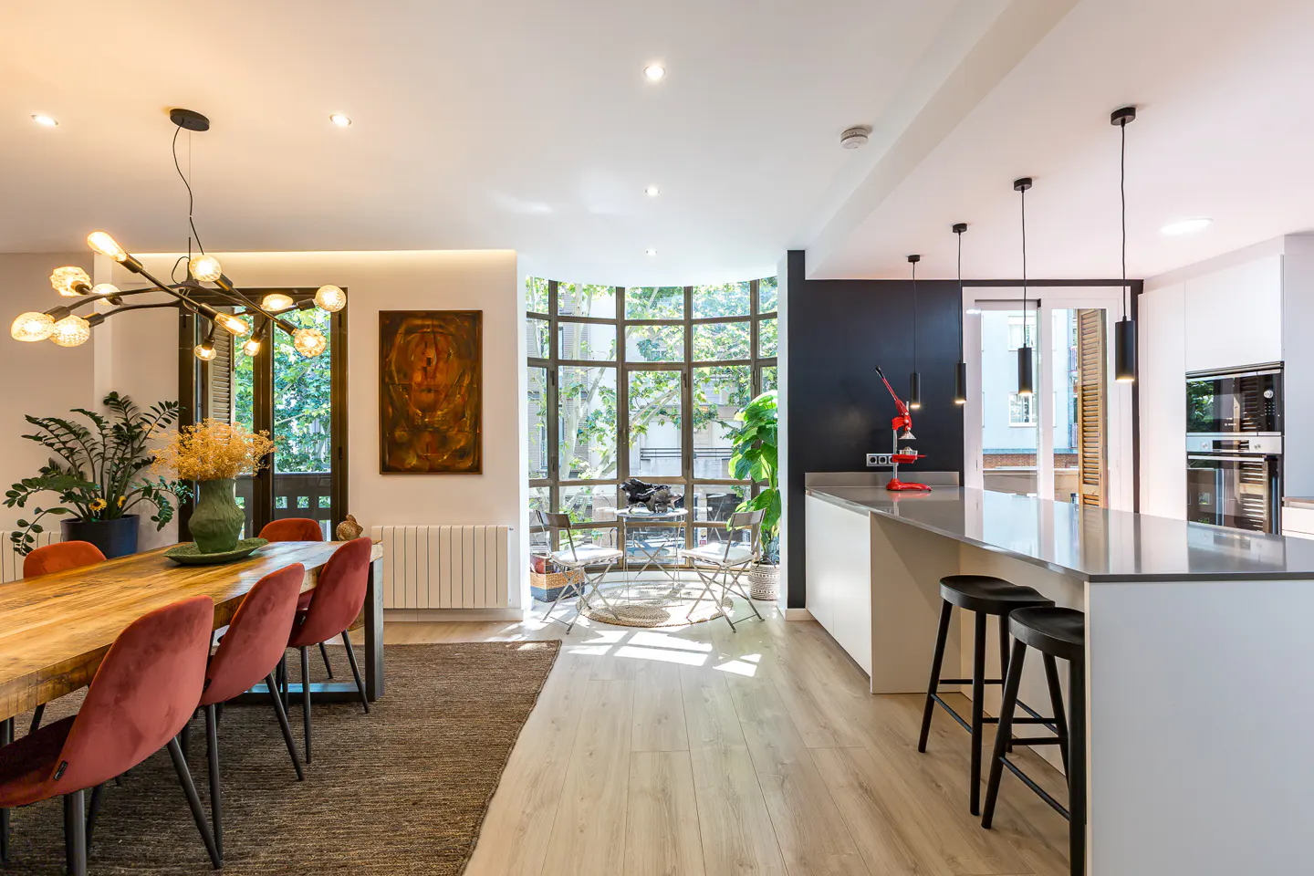 Open-concept dining and kitchen area with wood floors, a large window, and modern decor. Dining table with red velvet chairs. Kitchen island with black stools.