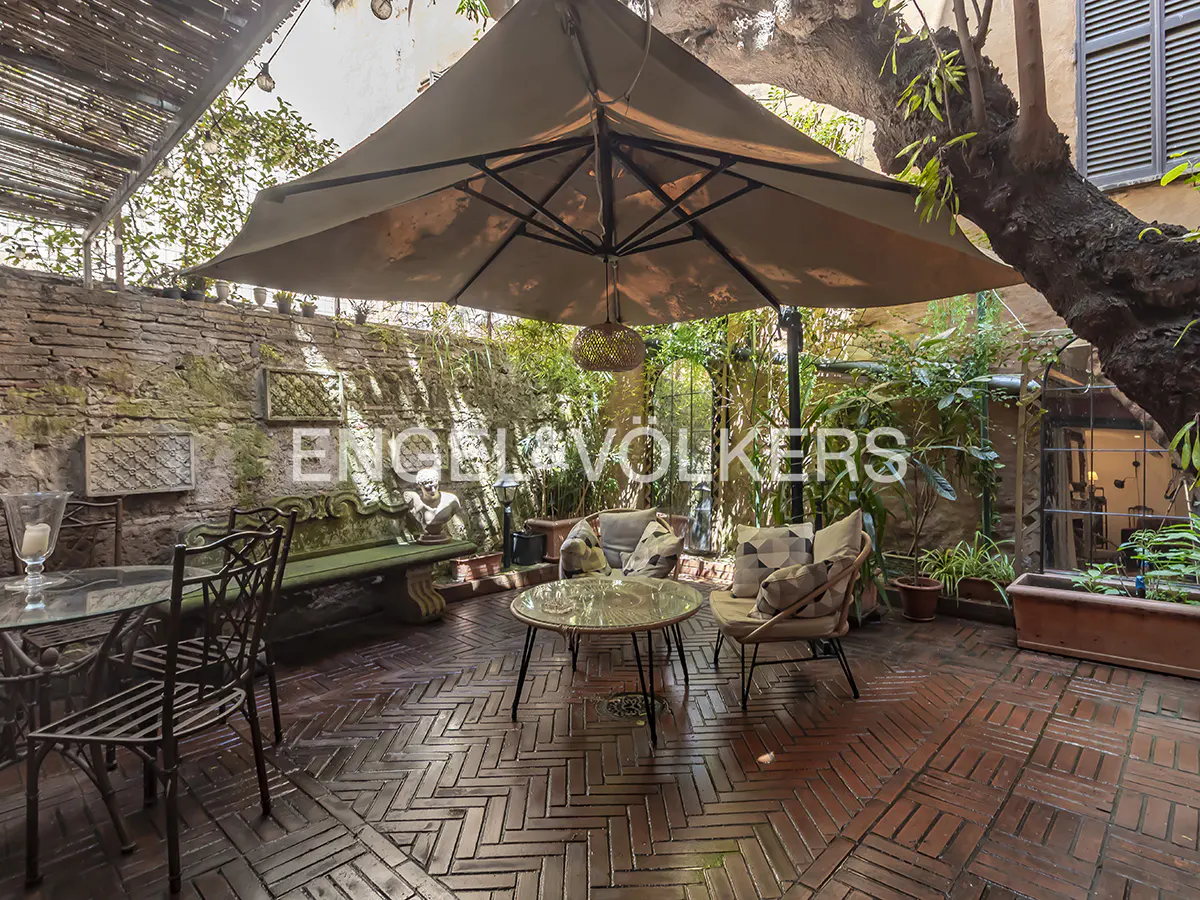 Outdoor patio with brick flooring, a large umbrella, and seating. Lush greenery surrounds the space, with a stone wall in the background.