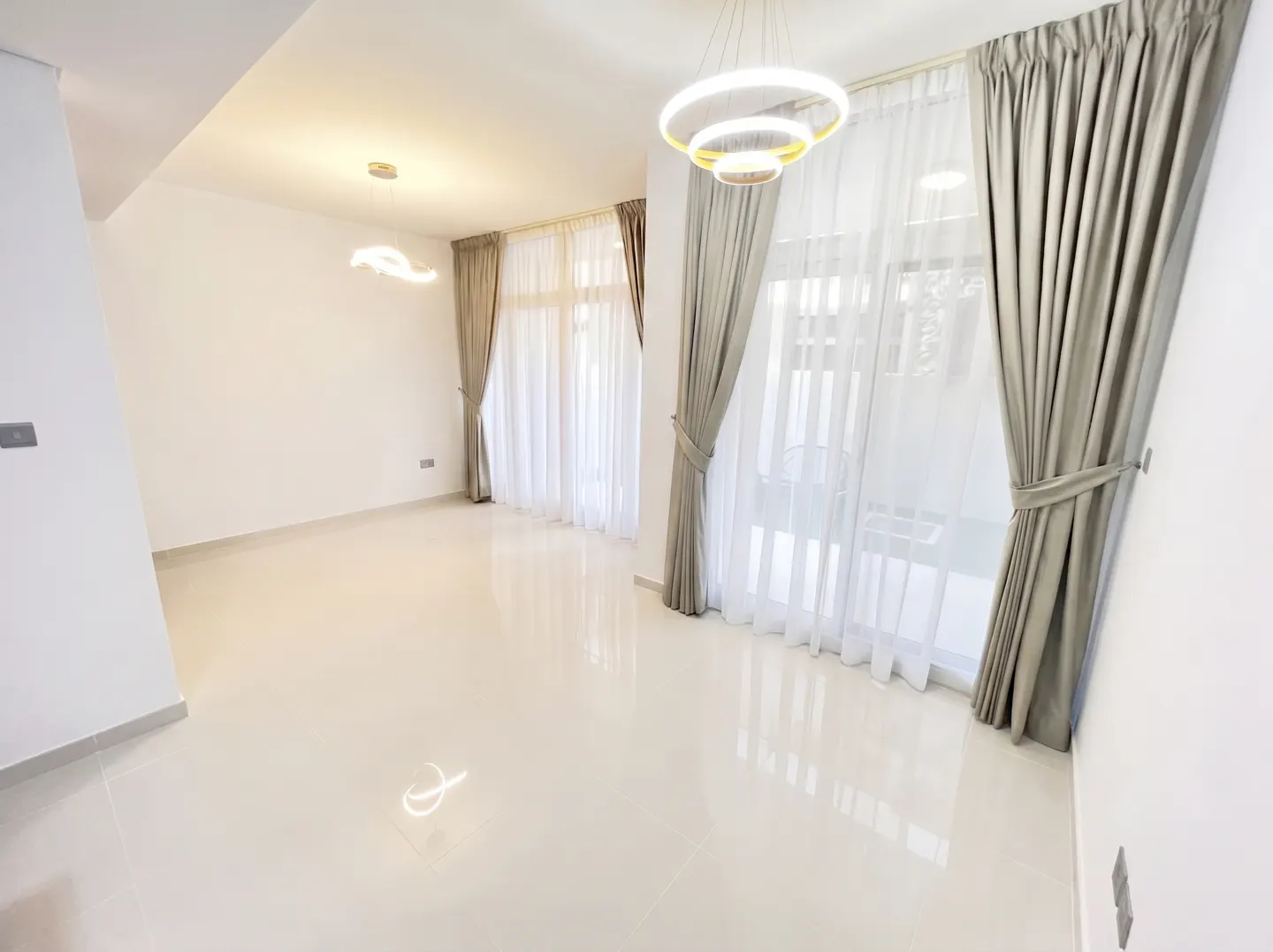 Bright, empty room with white walls and shiny tile floors. Windows have sheer white curtains and gray drapes. A modern, circular light fixture hangs from the ceiling.