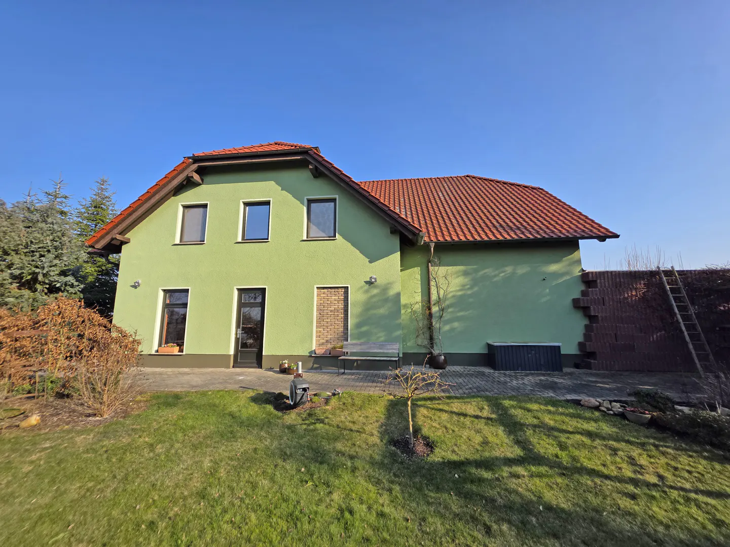 Two-story green house with a red tile roof, a brick wall under construction, and a ladder against the wall.
