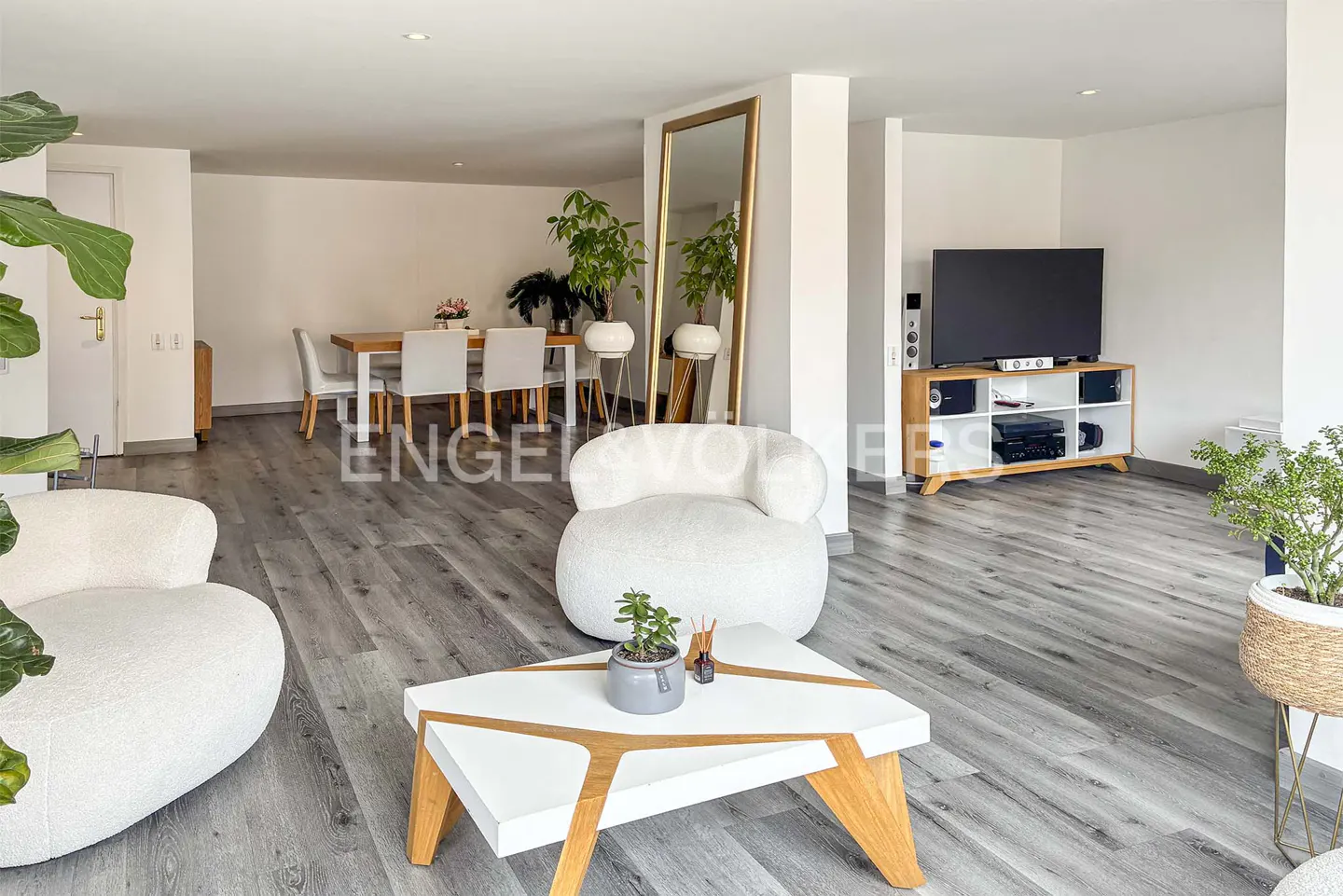 Bright, open-concept living room with white chairs, a modern coffee table, and a dining area in the background. A large mirror and TV are also visible.