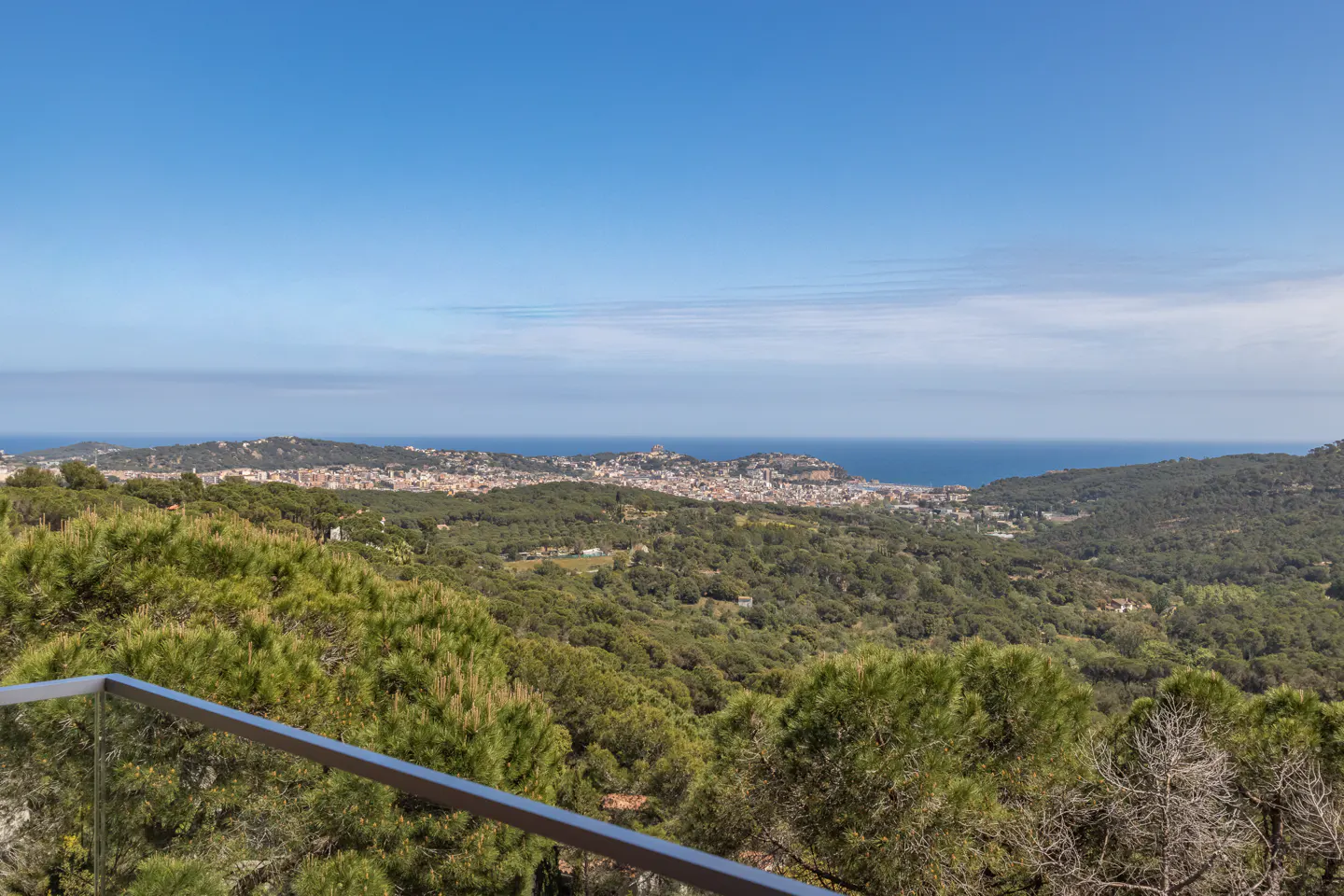Scenic view from a balcony with a metal railing, overlooking green hills, a coastal city, and the blue ocean under a clear sky.