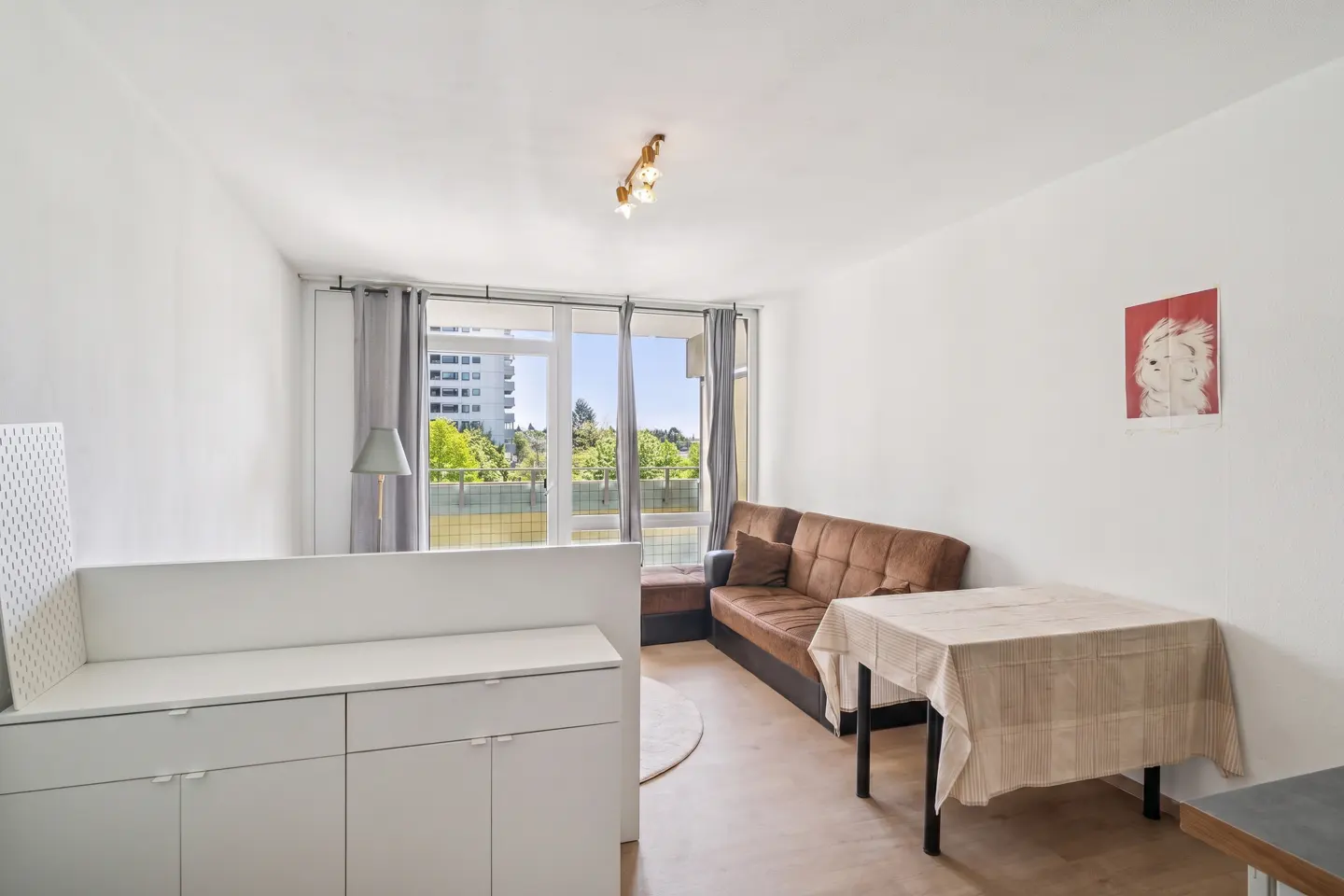 Bright apartment interior with white walls, brown sofa, and table with a patterned tablecloth. Balcony doors open to a view of trees and a building.