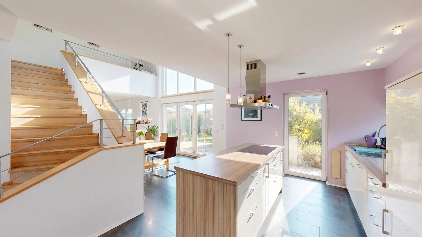 Bright, modern kitchen with wood stairs, island, and dining area. Large windows offer a view of the outdoors. The walls are a light purple.
