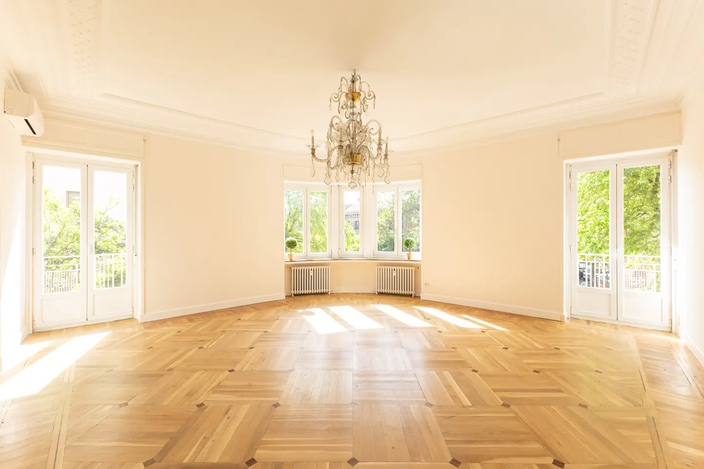 Bright, empty room with parquet floors, cream walls, and a chandelier. Windows and doors let in natural light.