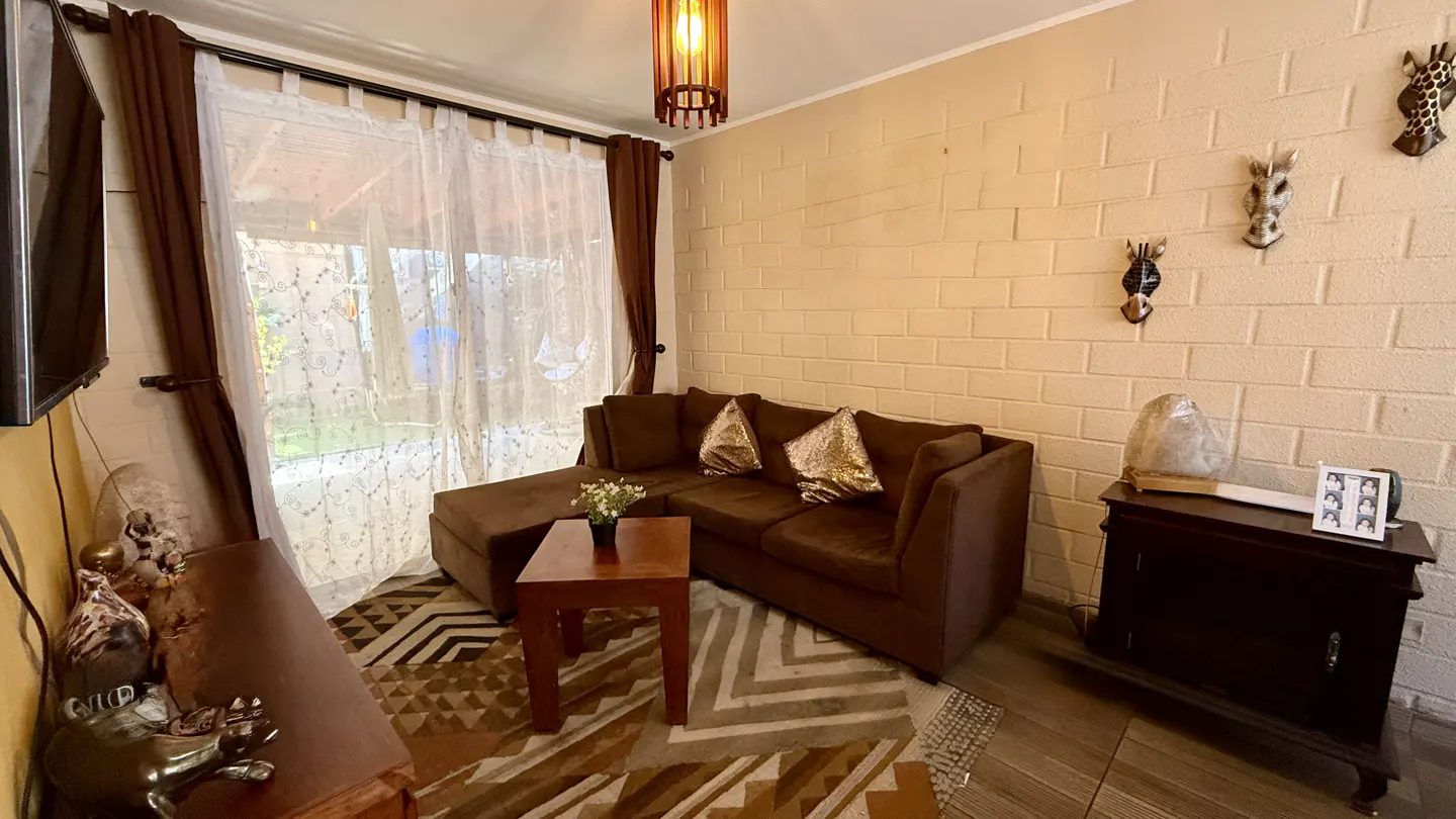 Living room with brown sectional sofa, wood table, and brick wall with animal masks. Curtains frame a bright window.
