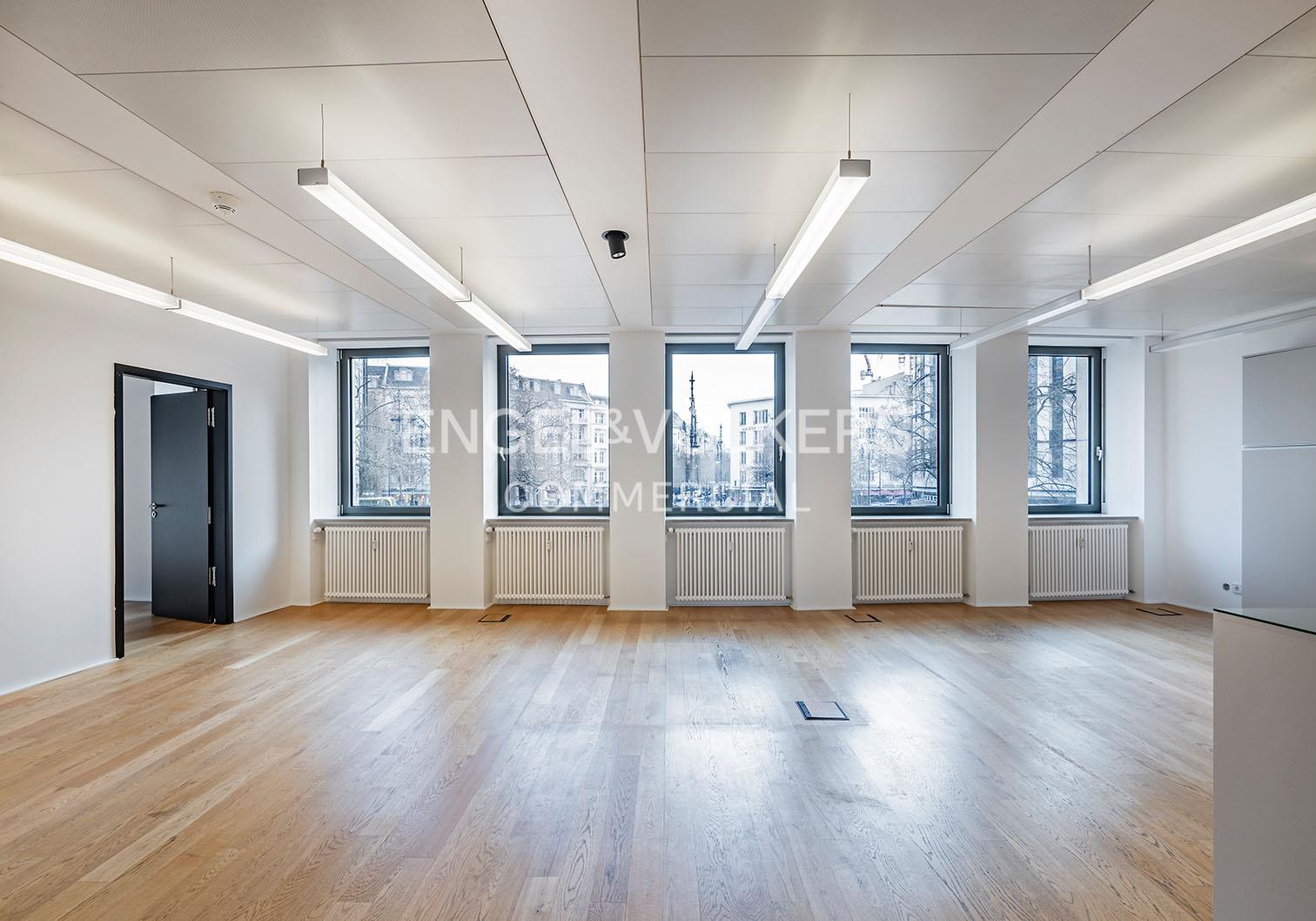 Bright, empty office space with light wood floors, white walls, linear ceiling lights, and large windows overlooking a city.
