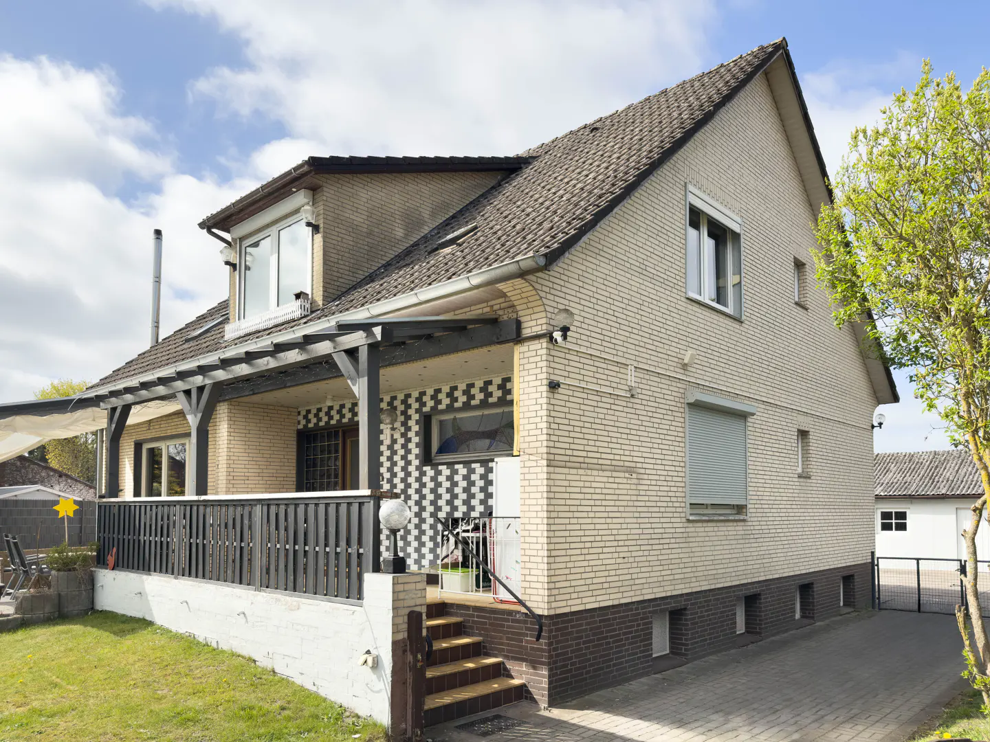 Two-story beige brick house with a dark brown roof and a covered porch with black railings. A paved driveway leads to a garage in the background.