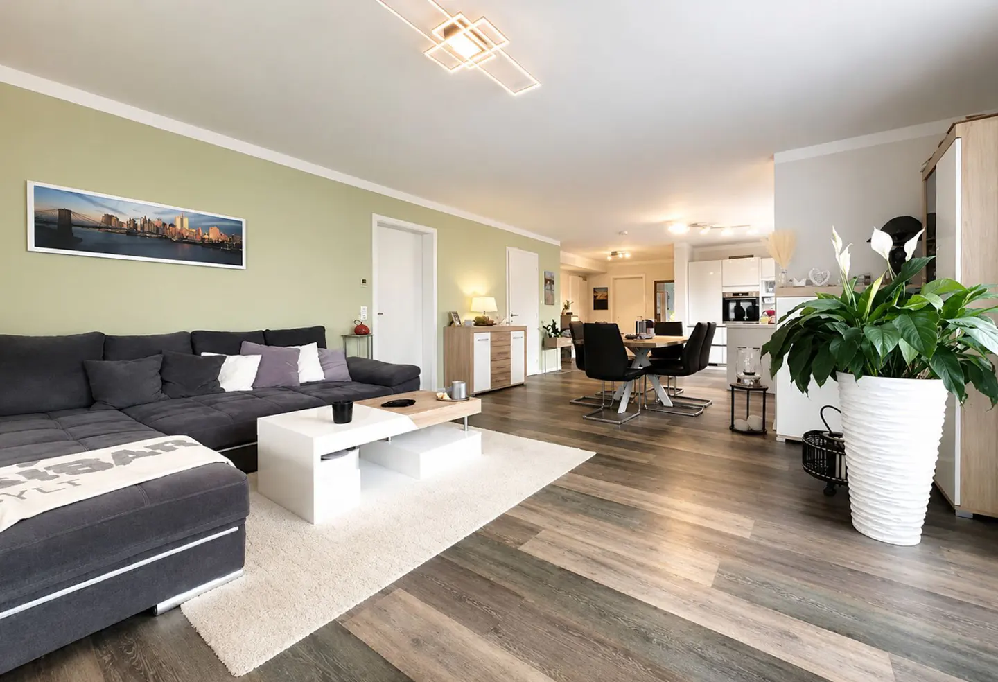 Modern open-plan living room with a grey sectional sofa, white coffee table, and dining area on wood-look flooring.