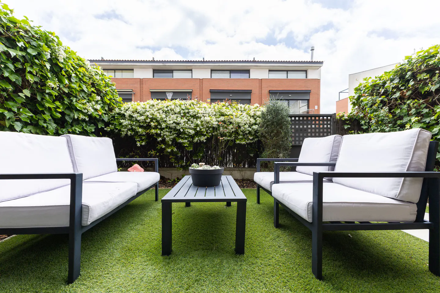Outdoor patio with two white sofas, a gray table, and green grass. A brick building is visible in the background.