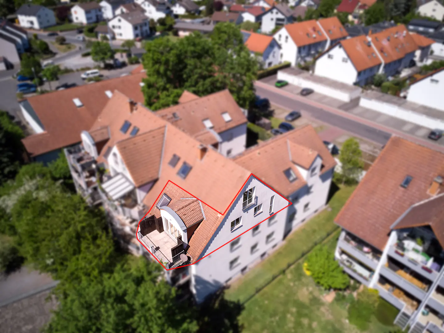 Aerial view of a multi-story apartment building with a red outline, featuring a balcony with chairs and a table. The building has a red tile roof and white walls.