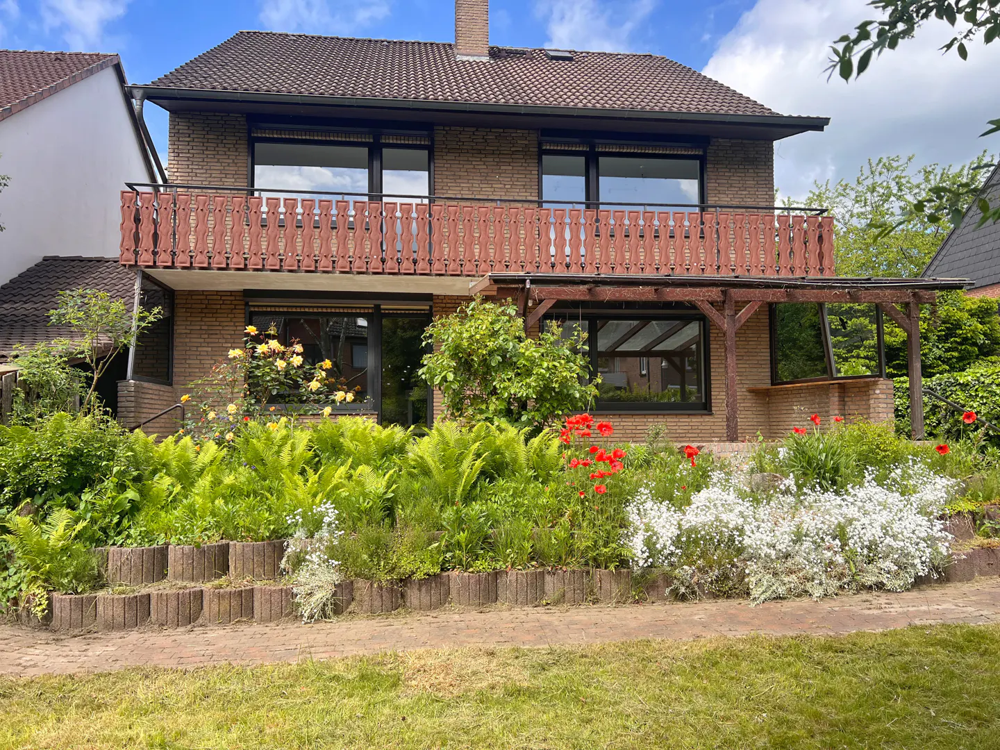 Two-story brick house with a brown roof, balcony, and a garden with red and white flowers.