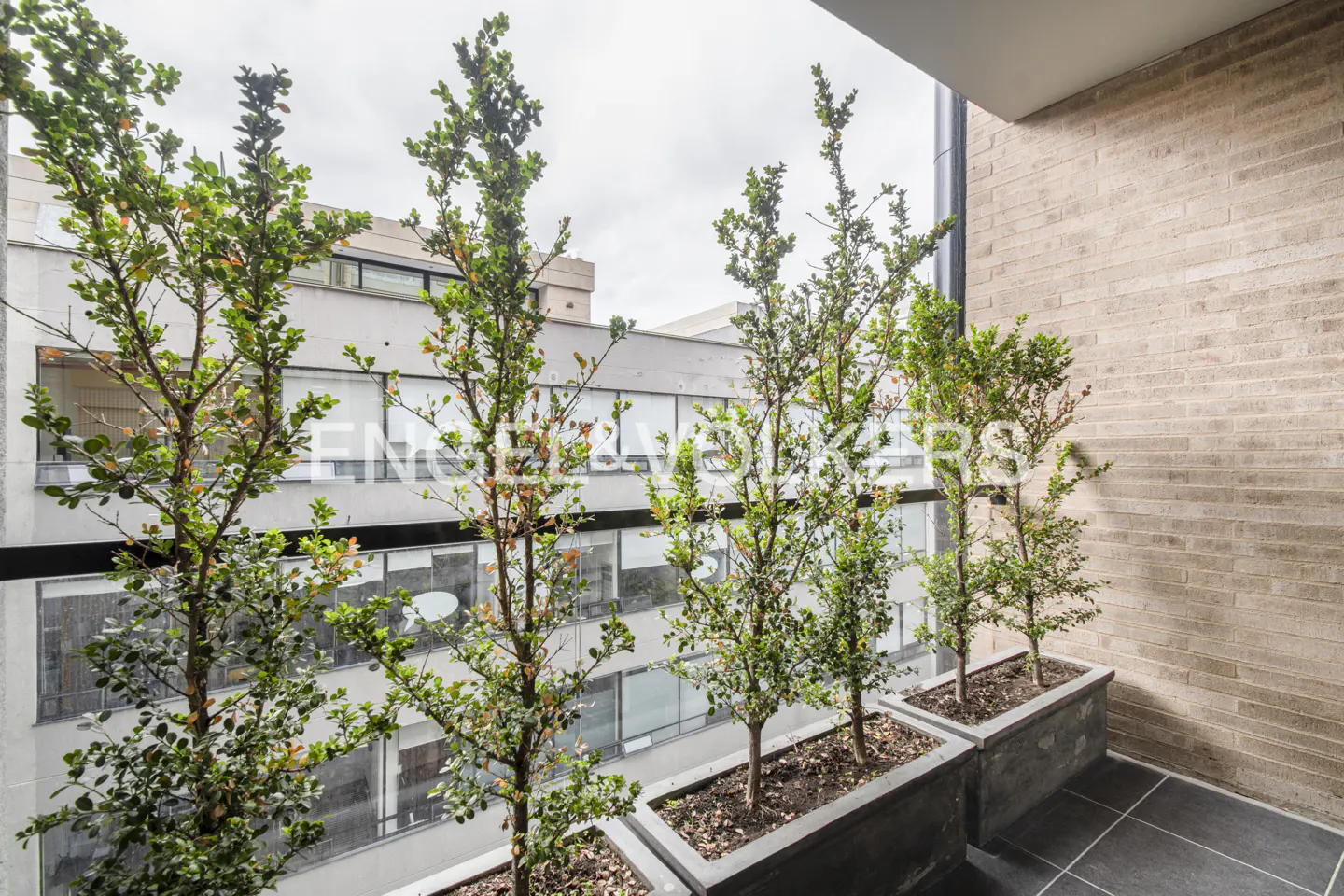 Balcony view with potted trees, gray tile floor, and brick wall. Building with windows visible through the trees. Overcast sky.