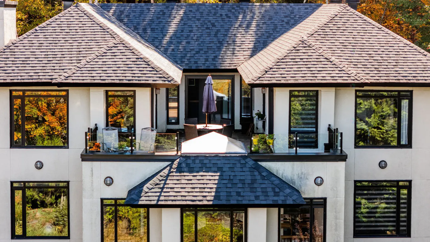 Close-up of a two-story house with a gray roof, white walls, and black-framed windows. A patio with an umbrella is visible on the second floor.