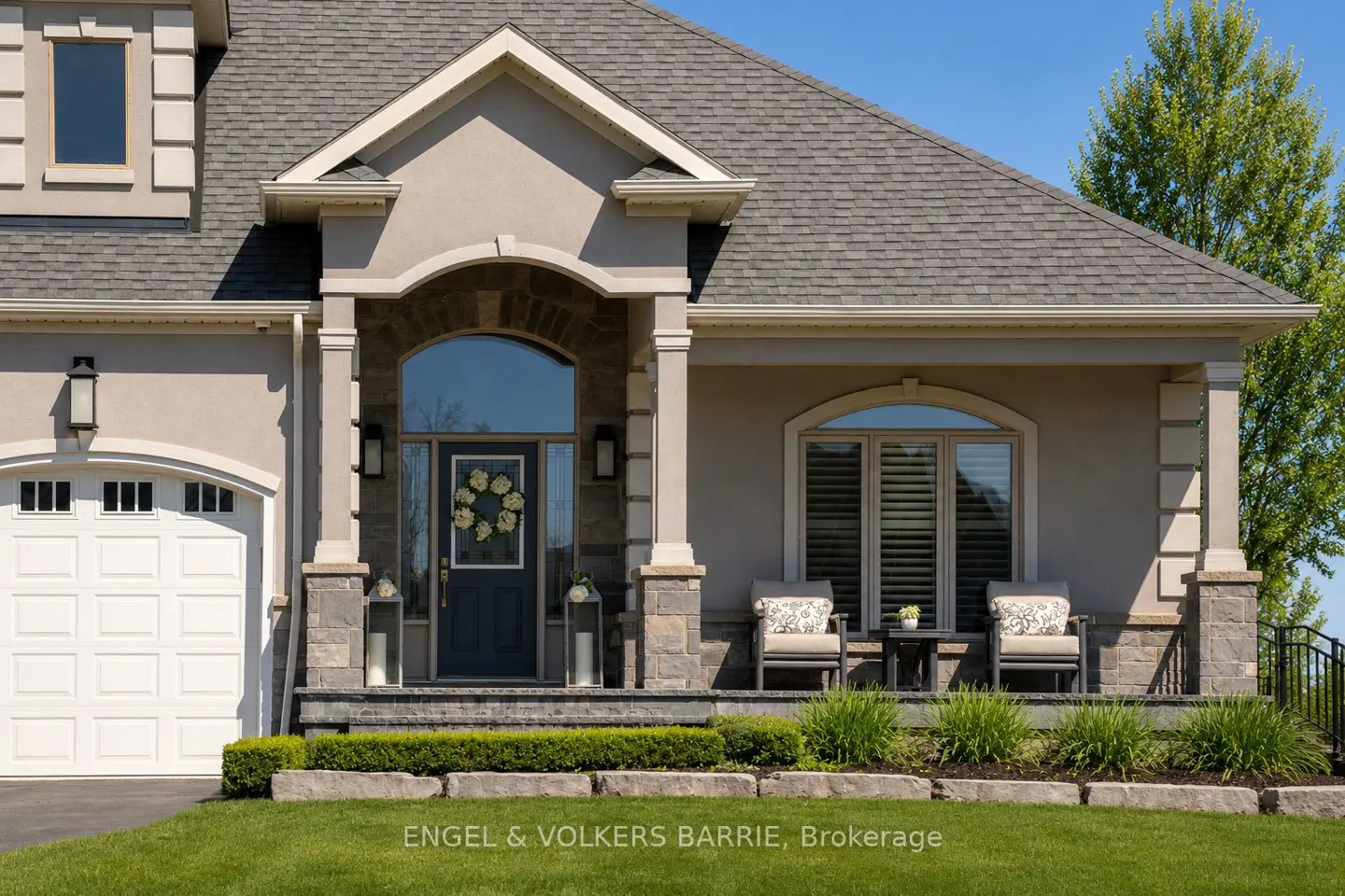 Beige house with gray roof, white garage door, and a blue front door with a wreath. Two chairs sit on the porch.