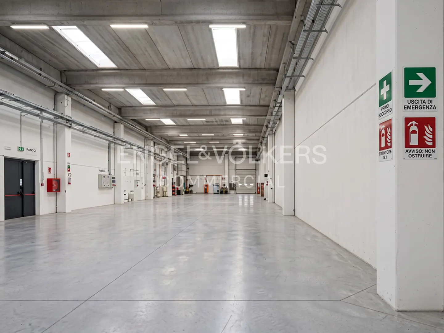 A bright, empty warehouse with a concrete floor and ceiling. Emergency exit signs are visible on the right wall.