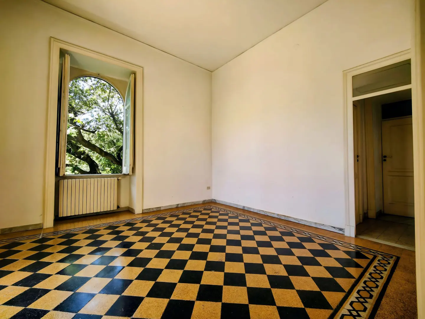 Empty room with black and gold checkered floor. An open window shows green trees. A white door is on the right.