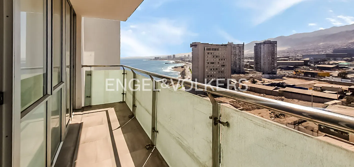 Balcony view of a coastal city with tall buildings, ocean, and mountains under a blue sky.