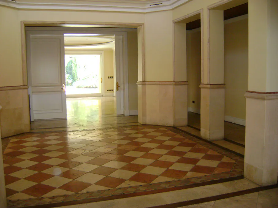 Interior view of a home with cream walls, columns, and a checkered tile floor. White double doors lead to a bright room with a large window.